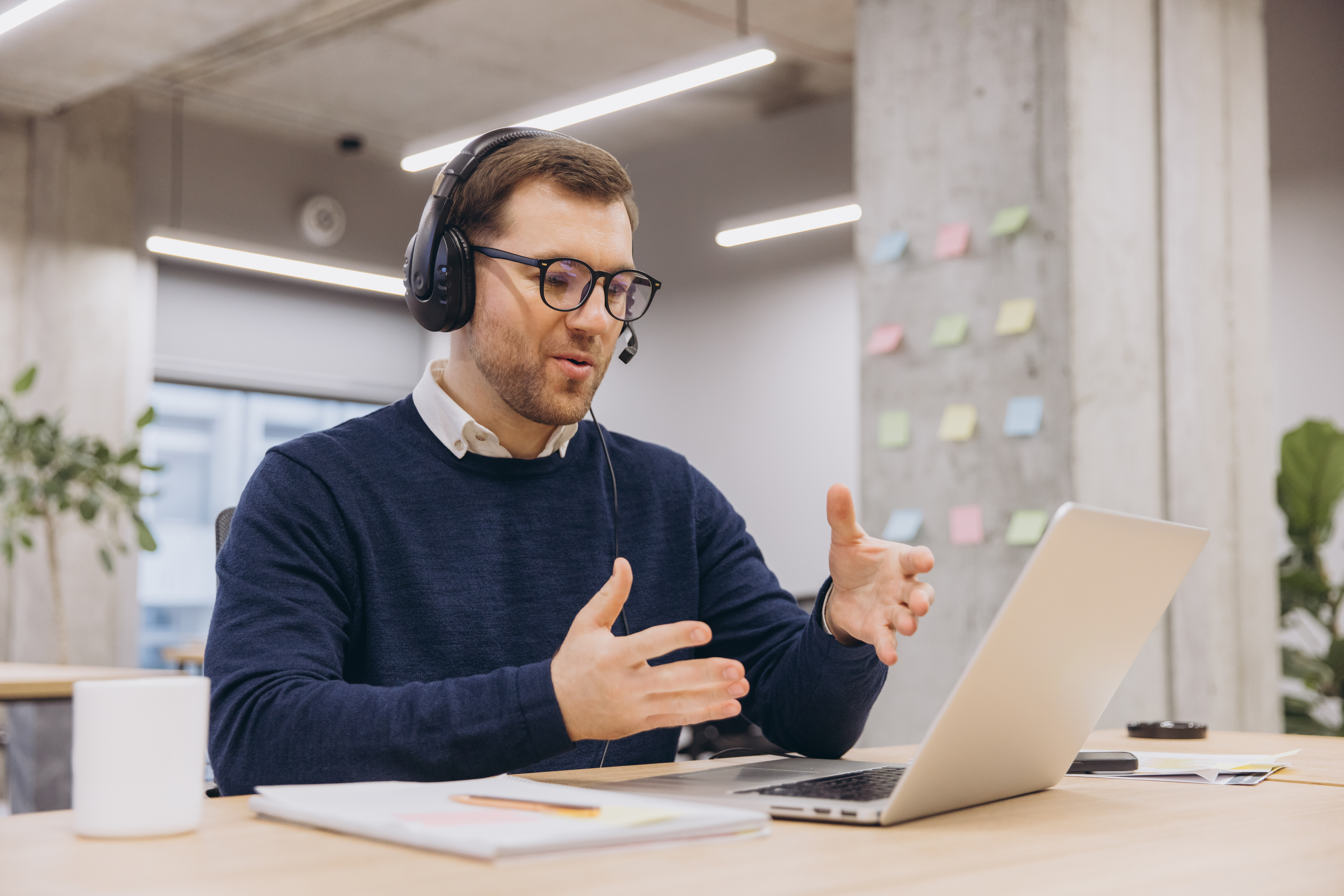 Office male worker wearing headset and glasses gesturing while having video conference on laptop in modern office