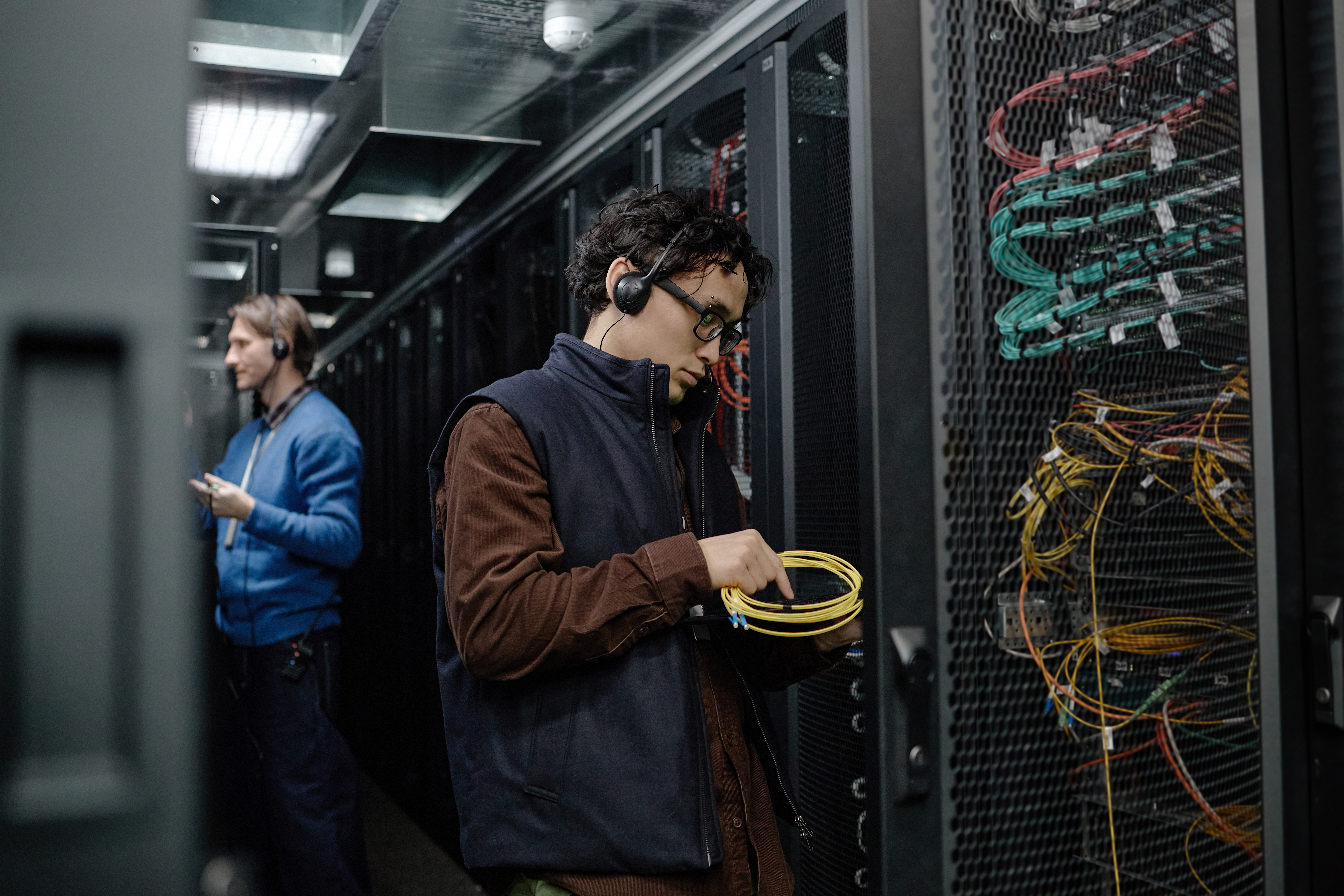 Young adult man wearing headset working with network cables in server room, middle aged Caucasian man in background checking data on digital tablet among server racks